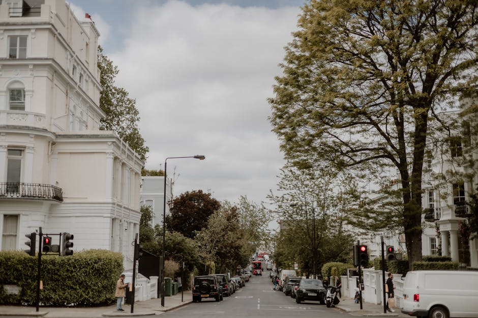 A quiet street in Hendon featuring white Victorian-style terraced houses with ornate detailing and wrought-iron balconies. Lush green trees line both sides of the road, with some leaves beginning to t