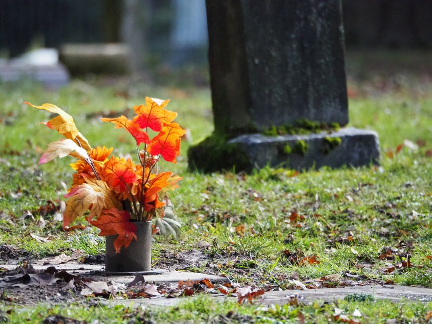 A floral arrangement of artificial autumn leaves in shades of orange, red, yellow, and beige placed inside a small, cylindrical, dark grey vase on a grassy area at a cemetery. The leaves are arranged