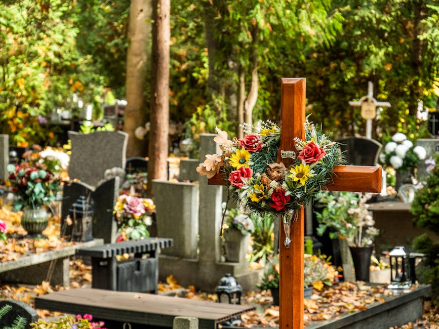A wooden memorial cross adorned with a floral arrangement featuring red roses, yellow daffodils, pink lilies, and greenery placed on a gravestone in a cemetery. The flowers appear fresh, with vibrant