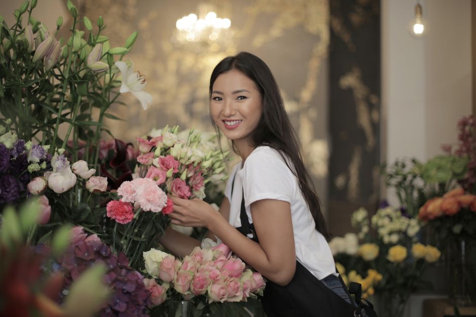 A young woman with long dark hair and a bright smile is selecting a bouquet of fresh flowers inside a floral shop. She is wearing a white T-shirt and black apron, holding a bunch of pink and white blo