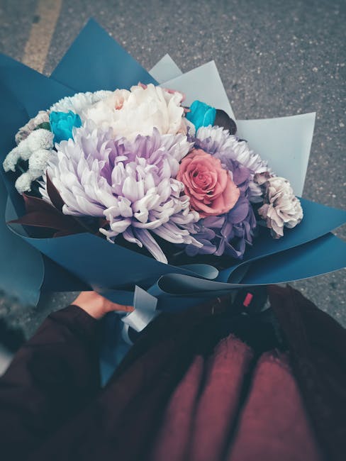 A close-up view of a floral bouquet featuring white, lavender, and pink chrysanthemums, along with a single pink rose and smaller purple and white flowers. The bouquet is wrapped in elegant blue and g