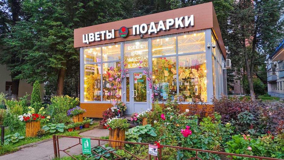A flower shop with large glass windows and a gray door, decorated with a floral wreath around the entrance, displaying a variety of fresh flowers inside. Outside, vibrant flower arrangements and potte